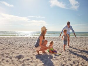 During their family summer vacation in Belize, a woman crouches next to a toddler playing with a sand toy as a man chases a running child. The ocean provides a stunning backdrop to their unforgettable day under the partly cloudy sky.