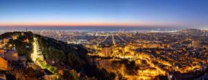 Panoramic view of Barcelona at twilight, capturing a vibrant European cityscape with illuminated streets and buildings. The horizon shows the sky transitioning from deep blue to orange as the sun sets behind the distant sea, making it one of the easiest ways to experience iconic holiday destinations.